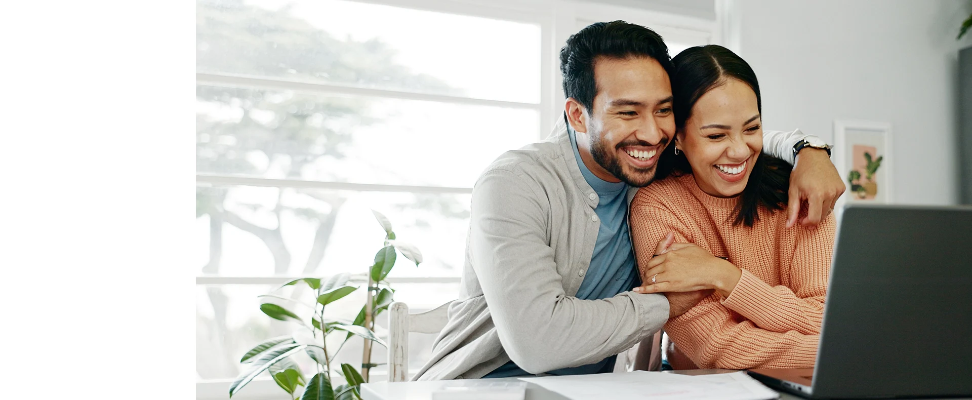 A couple sits close together at a table with laptops and a plant, sharing a cozy moment in a bright, modern workspace.